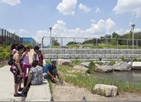 A group of students and their chaperones at a Hunts Point waterfront park pause at the water's edge as their instructor squats down to point out something the sand