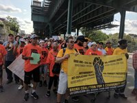 Large group of people marching with signs and banners in the streets underneath an elevated subway track.