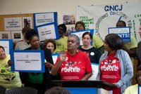 A group assembles behind a podium holding signs that spell out various demands while two women in matching tshirts address the crowd