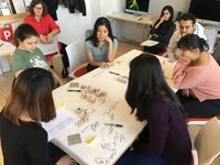 A group of young people sits around a table covered with paper and holding small wooden block objects and markers.