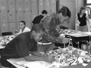 Young people stand around clusters of desks set up in a classroom while they work with small toys and objects