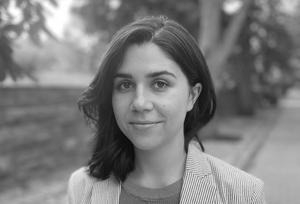 Black and white photo. Headshot. Young woman with dark hair in striped blazer, smiling, background is a park setting