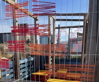 Red and orange colored string creates a weaved pattern in the foreground. In the background, a rooftop with colorful furniture.